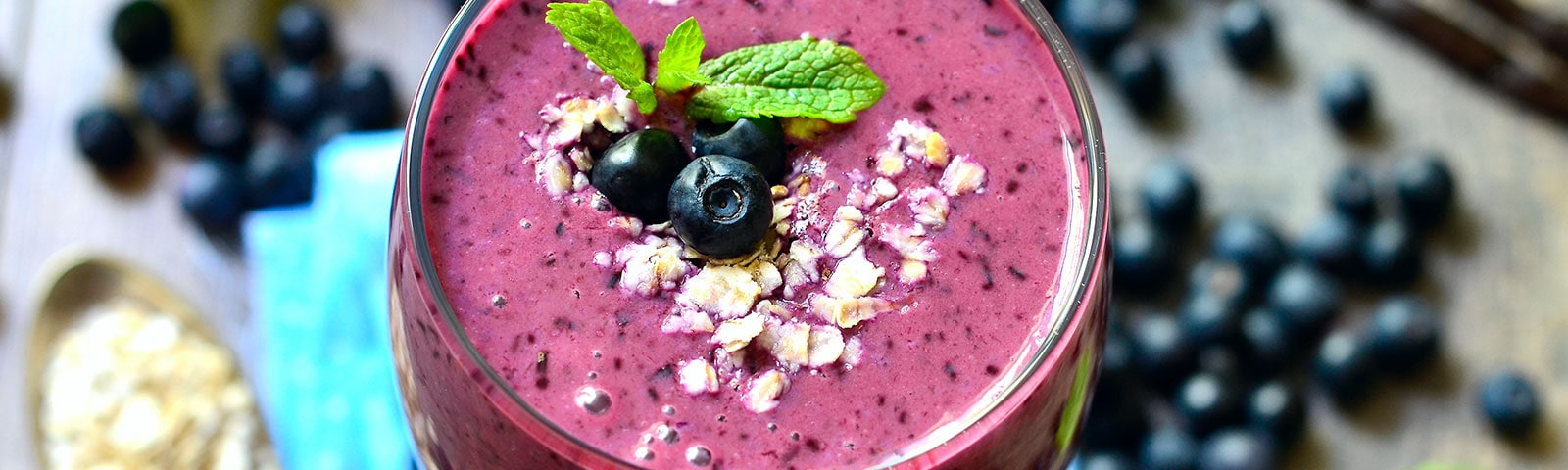 Center frame is a smoothie in a glass topped with blueberries and a mint leaf. Out of focus in the background blueberries and mint sit on a table.