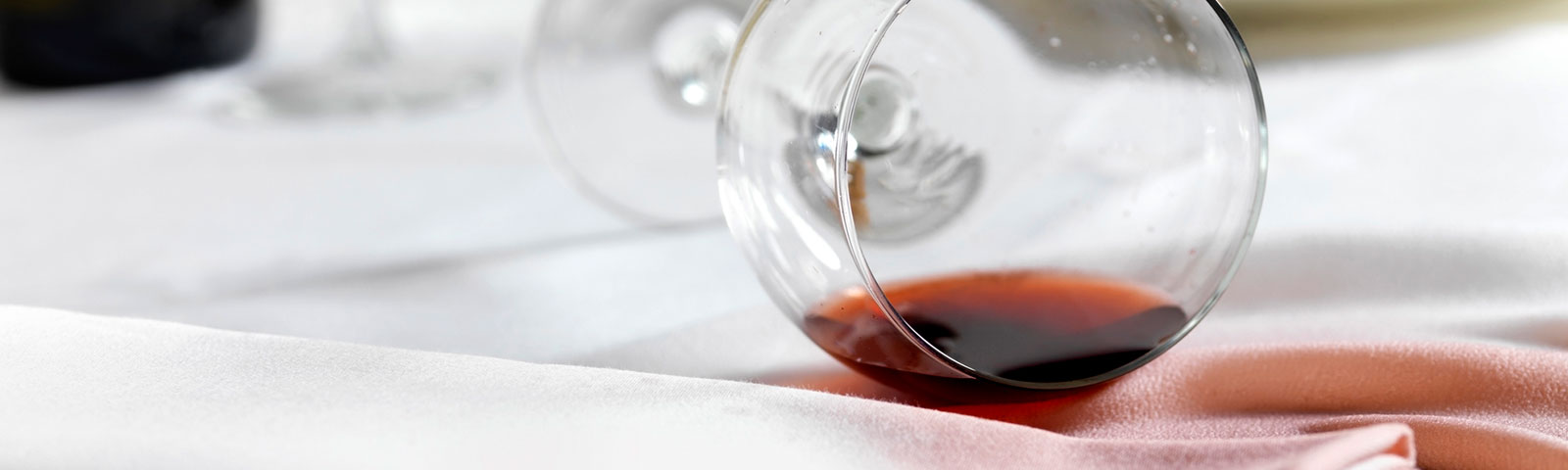 Wine glass on its side, spilling out its contents of red wine onto a white linen table cloth and creating a stain