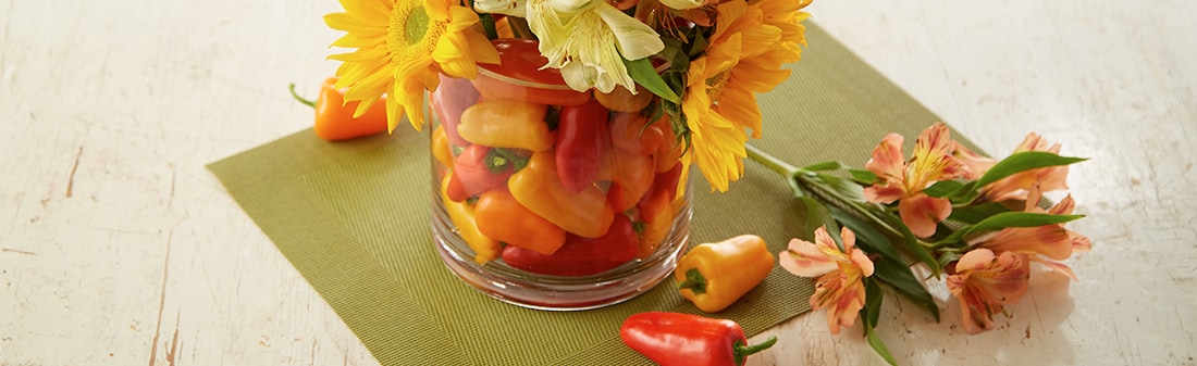 A vase full of multicolored mini peppers with sunflowers coming out of the top