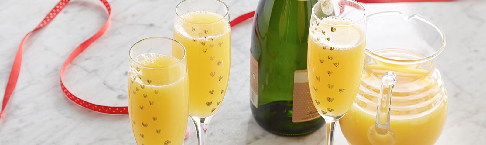 Crafty champagne flutes filled with mimosas in the foreground and candy hearts scattered on a marble countertop 
