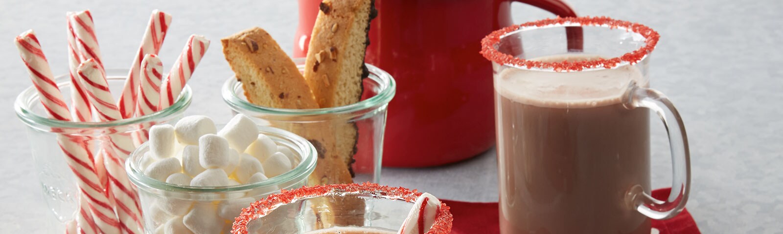 A cup of hot cocoa with a colorful red candied rim sits next to three cups containing candy cane sticks, biscotti and marshmallow respectively